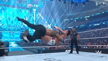 Two wrestlers are engaging in a dramatic move in a wrestling ring under bright blue lighting. The background features an enthusiastic crowd and a large 'WrestleMania' display.