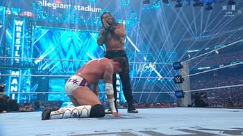 A dramatic scene from a wrestling match at WrestleMania, featuring a muscular wrestler smiling triumphantly over another who is kneeling in defeat. The background is illuminated with blue lights and the WrestleMania logo is displayed prominently.