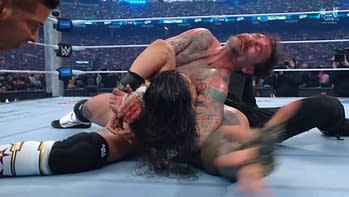 A dramatic wrestling scene shows two fighters in a grappling hold on the mat, with one wrestler displaying visible blood and tattoos. The background features a packed arena with bright lights, indicating an intense match atmosphere.