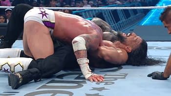 A wrestling match scene featuring two competitors, one on top of the other, with visible facial expressions of effort and pain. The arena is filled with spectators in the background.