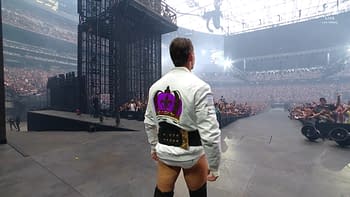 A wrestler, seen from behind, wearing a white jacket with a purple crown design and a championship belt, stands on a large stage in front of a packed stadium. The audience is cheering as bright lights illuminate the scene.