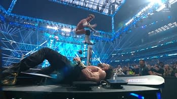 A wrestler performs a daring leap off the top rope while another wrestler lies prone on the ground, surrounded by a vibrant arena filled with blue lights and an enthusiastic crowd. The scene captures the intensity of a wrestling match.