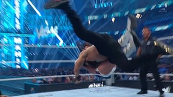 A wrestler performs a dynamic move during a high-energy match in a wrestling ring, with a vibrant blue lighting setup in the background.