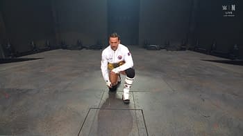 A professional wrestler kneels on a stage, wearing a white jacket and black trunks, with a championship belt secured around his waist. The background features a dark arena setting with lights and seating in the distance.