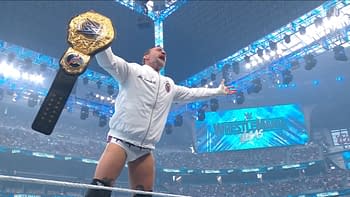 A wrestler celebrates in the ring, holding a championship belt above his head. The background features bright lights and a large screen displaying 'WrestleMania.'
