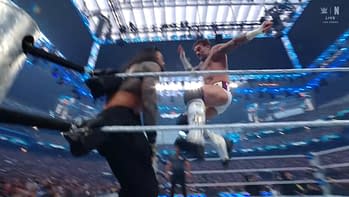 A wrestler in mid-air performs a kick while another wrestler, with long hair, stands nearby, looking up. The scene captures the energy of a live wrestling match. Bright arena lights illuminate the action.