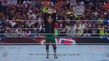 A wrestler stands triumphantly in a ring, holding up a championship belt. The crowd behind him is animated and holds various signs, creating a lively atmosphere.