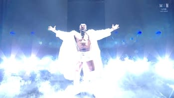 A wrestler poses dramatically on stage, wearing a white fur coat and shorts, with arms outstretched as bright lights and smoke surround him.