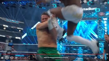 A wrestler jumps in the air with one leg extended while another wrestler, holding his leg, braces for impact. The stadium is illuminated with bright blue lights, and the audience can be seen in the background.