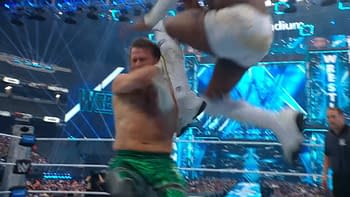 A professional wrestling match scene featuring one wrestler performing a jump maneuver towards another wrestler, who is bracing for impact. The background shows a vibrant arena with bright lights and a WrestleMania banner.