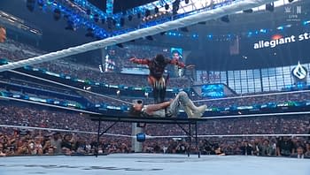 A wrestler performs a high-flying move from the ropes onto an opponent lying on a table in a packed stadium during a wrestling event. The audience is captivated by the action, with fans cheering enthusiastically in the background.