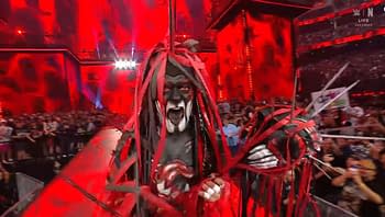 A wrestler with dark face paint and elaborate red and black costume filled with ribbons and spikes stands in a ring surrounded by fans. The arena is lit with dramatic red lighting, creating an intense atmosphere.