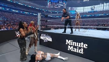 WrestleMania scene featuring Rhea Ripley lying on the mat, while several other wrestlers react nearby. The crowd fills the stadium in the background.