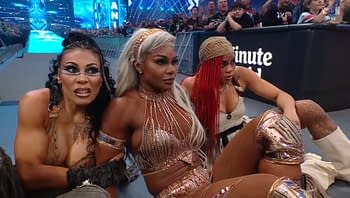 Three female wrestlers sit on the ground at WrestleMania, showcasing diverse hairstyles and outfits, with one having metallic accessories. They display expressions of concern or intrigue while watching the match.