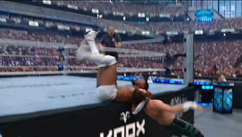 A wrestler performs a high-impact move off the ropes, airborne as he approaches another wrestler outside the ring, with a large crowd visible in the background at a wrestling event.