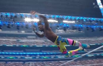 A muscular wrestler performs a high-flying move during a match, soaring through the air above a large crowd. He is wearing vibrant, colorful pants with patterns, and the arena is brightly lit with blue and white lights.