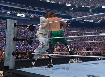 Two professional wrestlers are engaged in a match inside a crowded stadium, with one executing a move on the other. The audience is visible in the background, filled with fans watching the event.