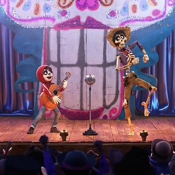 A scene from Coco featuring a young boy with a guitar and a skeleton in a hat performing on stage, with a colorful skull backdrop and an audience in front.