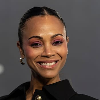 Zoe Saldana smiles at a premiere event, showcasing her striking makeup with purples and pinks. She wears a black outfit and hoop earrings, exuding confidence against a gray backdrop.
