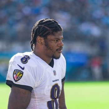 A focused Lamar Jackson, wearing a white Baltimore Ravens jersey with the number 8, walking on the field. The background is a blurred stadium filled with fans, indicating an NFL game atmosphere.