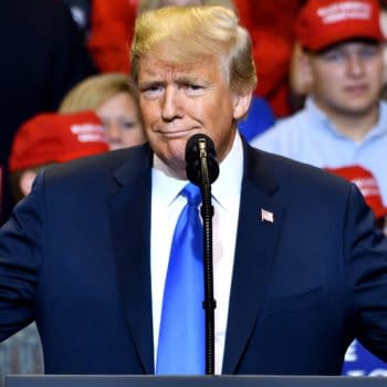 Donald Trump speaks at a campaign rally with a microphone in front of him, wearing a dark suit and blue tie. Supporters in red hats stand blurred in the background.