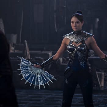 A female character from Mortal Kombat 2 stands ready for battle, wearing a metallic outfit with intricate designs and holding a pair of bladed fans. The dark and atmospheric setting adds intensity to the scene.