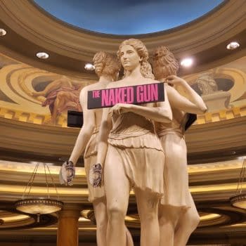 A trio of classical statues depicting women holding an oversized sign that reads 'The Naked Gun.' The background features ornate decorations and lighting, enhancing the setting.