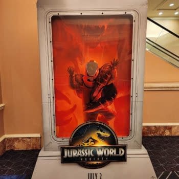 A promotional stand from 'Jurassic World: Rebirth' features a figure in a spacesuit behind a glass enclosure, surrounded by bright orange lighting that evokes a prehistoric atmosphere.