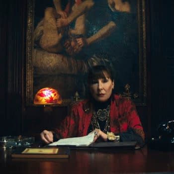 Anjelica Huston portrays The Director in a scene from 'Ballerina', seated at a desk in a richly decorated room filled with books and antiques. A dimly lit stained glass lamp and various objects adorn the desk, creating a dramatic atmosphere.