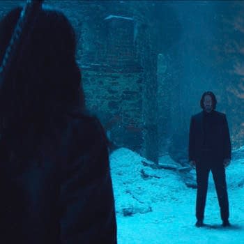 In a snowy, dimly lit setting, Keanu Reeves as John Wick stands face-to-face with an unseen character, creating a tense atmosphere in the film "Ballerina." The abandoned surroundings include partially collapsed walls and snow-covered ground.