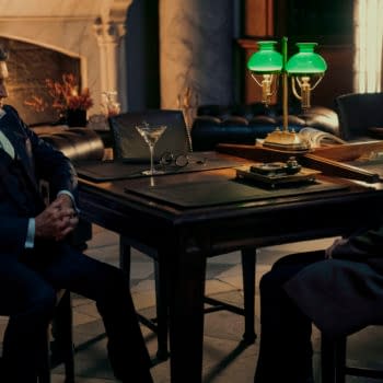 In a dimly lit room, Gabriel Byrne as The Chancellor sits across the table from Ana de Armas as Eve, both looking serious. A green lamp and a glass of martini add to the dramatic atmosphere.