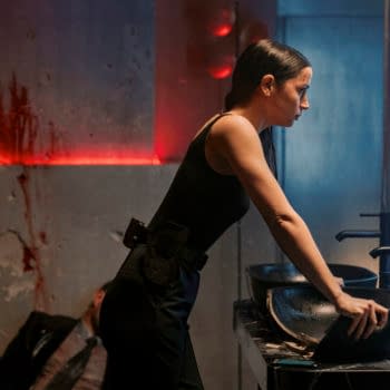 A woman in a black outfit leans over a black sink, her expression serious, in a dimly lit location with red accents and visible signs of distress. The scene hints at a tense moment in the film 'Ballerina,' featuring Ana de Armas as the character Eve.