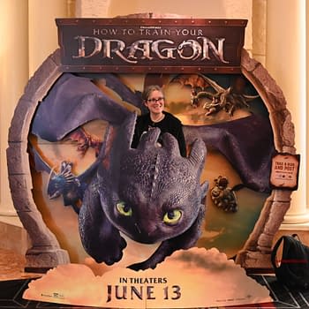 A person stands inside a promotional cutout featuring the dragon Toothless from 'How to Train Your Dragon', surrounded by colorful artwork of other dragons and characters. The cutout displays the release date, June 13, prominently at the bottom.