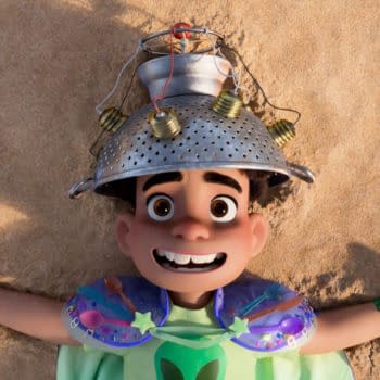 A scene from the animated film 'Elio' featuring a young boy with a cheerful expression lying on sandy ground, wearing a colander as a hat and colorful armor, embracing his imaginative and adventurous spirit. The film, produced by Disney and Pixar, explores themes of space and self-discovery.