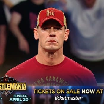 John Cena stands in the ring during WWE Raw in Brussels, wearing a red shirt and a red cap. The background shows blurred fans cheering.