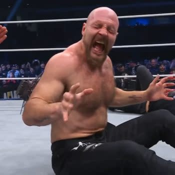 Jon Moxley, visibly in pain, sits in the wrestling ring with spikes protruding from his back after a confrontation with Adam Copeland. The referee is present, appearing to call for assistance.