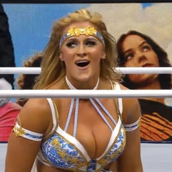 A wrestler, wearing a striking blue and gold outfit, expresses excitement after pinning her opponent during a match. The background shows a fan reacting to the action.
