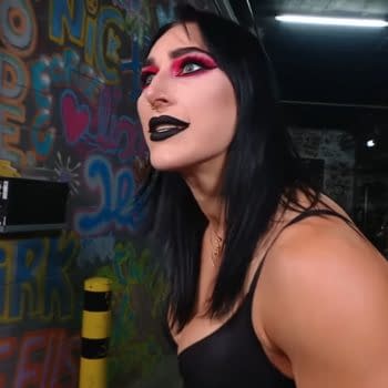 Rhea Ripley, a professional wrestler, is shown backstage at WWE Raw in Brussels, wearing striking makeup with bold pink and black colors. The background features colorful graffiti art, adding to the vibrant setting.