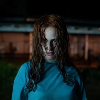 A woman with disheveled hair and an intense expression stands in a dimly lit outdoor setting, wearing a blue shirt. A small wound is visible on her forehead, adding to the eerie atmosphere.