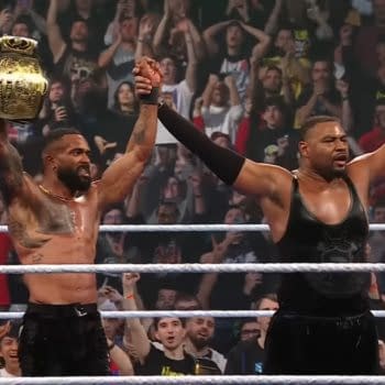 Two wrestlers from the Street Profits celebrate in the ring after winning a championship, with fans cheering in the background. One holds a championship belt aloft while the other raises his arm triumphantly.