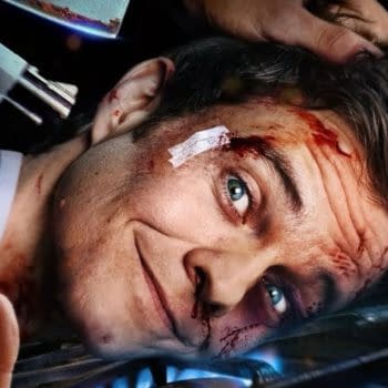 A man with a gruesome injury on his forehead and blood on his face is smiling, while a hand appears to be holding a gun near his head. Novocaine Review: A captivating exploration of chaos and humor amidst dark themes.