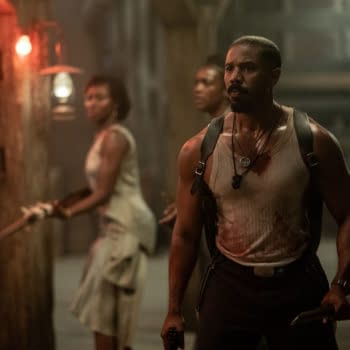 A tense scene from the movie 'Sinners', featuring a muscular man in a blood-stained sleeveless shirt holding a weapon, with two others in the background prepared for confrontation, illuminated by dim lighting.