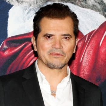 John Leguizamo poses on the red carpet at a movie premiere event in Hollywood, with a festive background featuring a Santa Claus image. He is dressed in a black suit and white shirt.