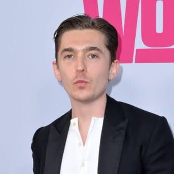 A man with short brown hair and earrings poses confidently in front of a light background featuring bright pink text, indicating a promotional event or premiere.