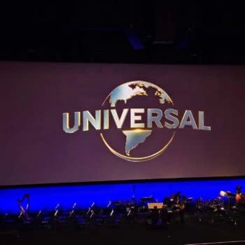 The Universal Pictures logo featuring a gold globe and the word 'UNIVERSAL' in bold letters appears on a large screen at a presentation event. In the foreground, there are music stands and instruments set up on stage.