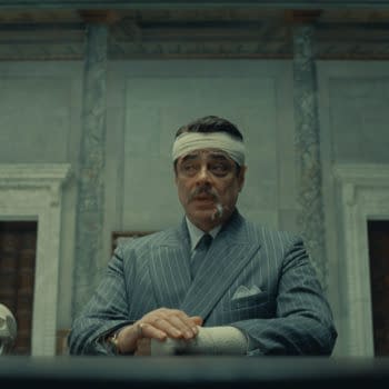 A scene from 'The Phoenician Scheme' featuring Benicio Del Toro as Zsa-Zsa Korda, dressed in a pinstripe suit with bandages on his head and hand, sitting at a table with a skull beside him in a richly decorated room.