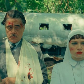 A still from the film "The Phoenician Scheme" featuring Benicio Del Toro as Zsa-Zsa Korda, wearing a blood-stained bandage, alongside Mia Threapleton as Liesl, who is dressed in a white outfit and a headscarf. The background shows a damaged airplane amid lush greenery.