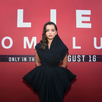 A woman in a dramatic black gown poses in front of a promotional backdrop featuring the title 'ALIEN: ROMULUS'. The backdrop is bright red, emphasizing the film release date.