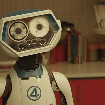 A friendly robot character named H.E.R.B.I.E stands in a modern kitchen setting, featuring a bright red cabinetry and various kitchen items in the background. The design includes a distinctive blue and white color scheme with the number '4' displayed, representing its connection to Marvel's 'The Fantastic Four: First Steps'.
