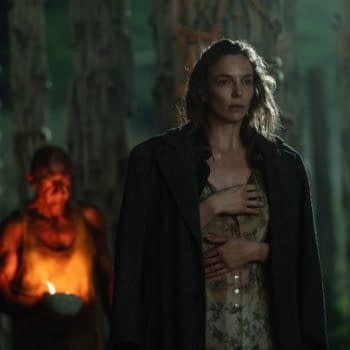 A dramatic scene featuring a woman with tousled hair wearing a floral dress and a long coat, holding her hand over her chest in a forest setting. In the background, a shadowy figure holds a lit candle, adding an eerie atmosphere.